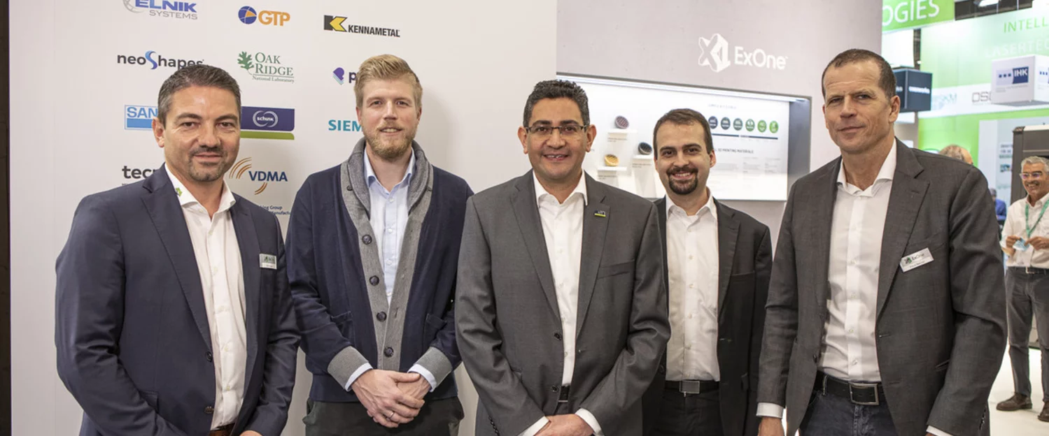 5 Mitarbeiter von Schunk und ExOne stehen vor einem Messestand