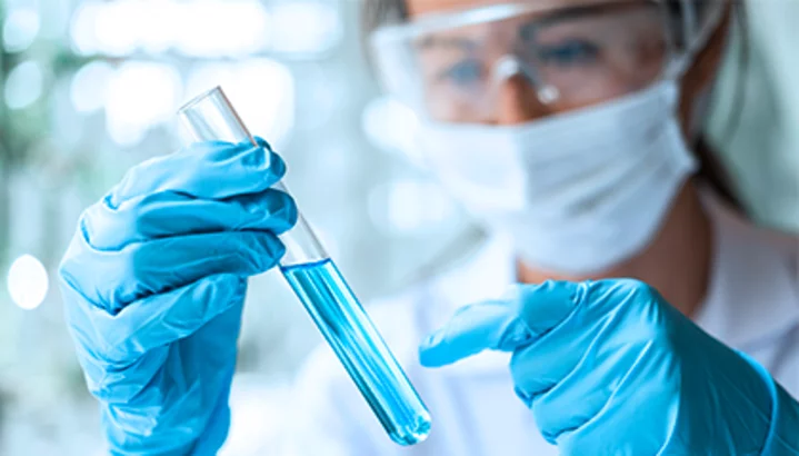 Lab technician in protective gear holding a test tube with blue liquid in a clinical laboratory setting.